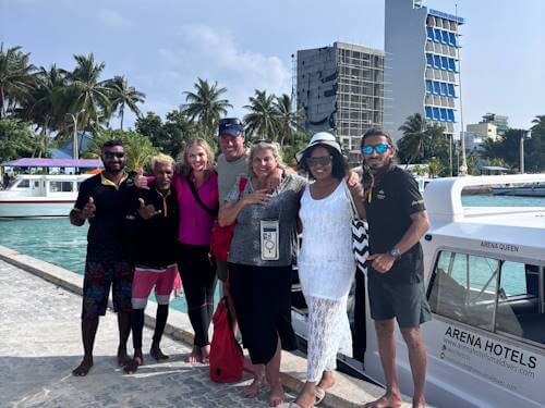 Happy group of tourists posing barefoot at a jetty near an Arena Hotels speedboat in the Maldives, with palm trees and buildings in the background