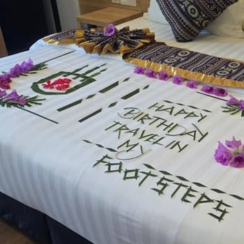 Tropical birthday message made with leaves and flowers on a hotel bed, reading "Happy Birthday Travel in My Footsteps" with a flower-decorated cake symbol