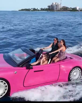 Four women riding a pink convertible-shaped boat on the open sea with city skyline and palm trees in the background